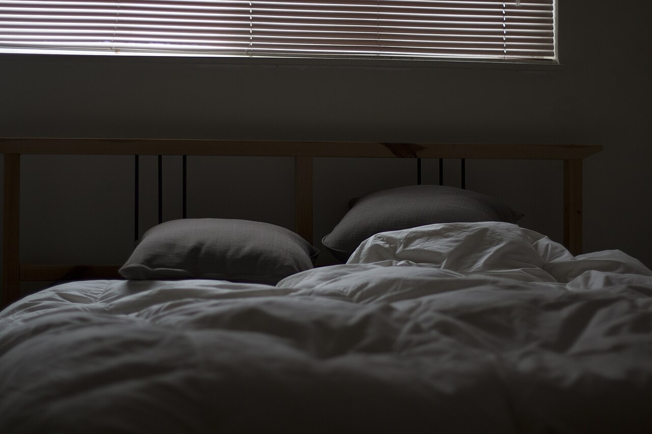 Morning light coming through closed venetian blinds into a bedroom, the bed unmade